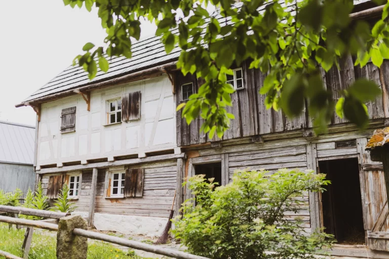 Fachwerkhaus mit Holzfassade und Fensterläden, im Vordergrund Laubzweige und Gartenzaun