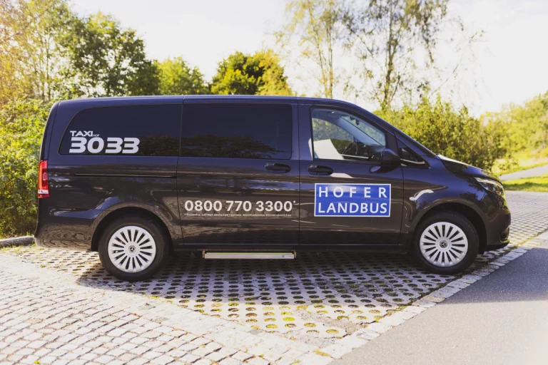 Dunkler Kleinbus mit Beschriftung „Hofer Landbus“ und Taxi-Rufnummer parkt auf gepflasterter Fläche