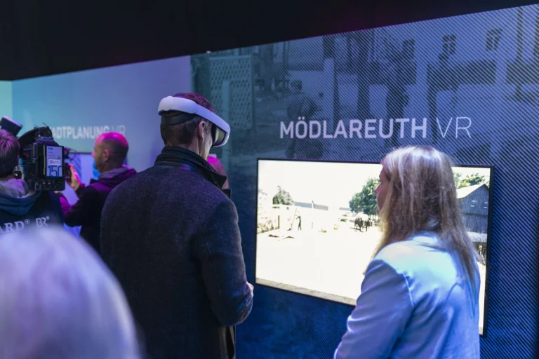 Person mit VR-Headset und weitere Personen vor einem Bildschirm mit der Anzeige „Mödlareuth VR“