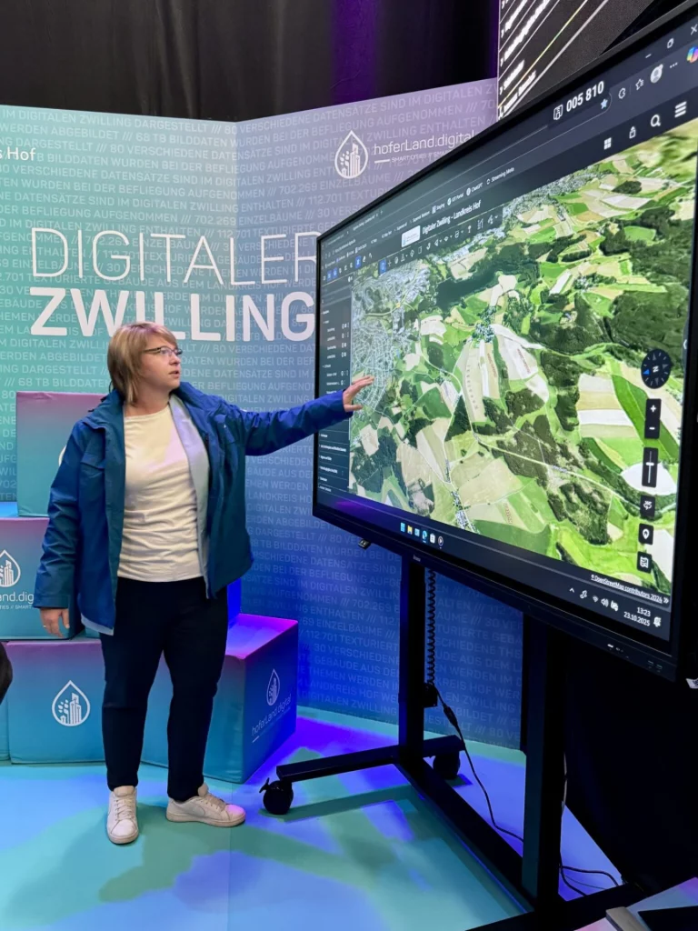 Frau erklärt digitalen Zwilling des Hofer Lands auf großem Bildschirm mit Luftbildkarte