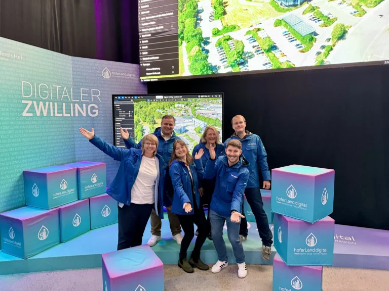 Gruppe von Personen in blauen Jacken vor Ausstellungswand „Digitaler Zwilling“ mit hoferLand.digital-Würfeln