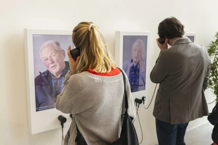 Besucherin und Besucher hören an Porträts mit Telefonhörern Zeitzeugenberichte in der Ausstellung