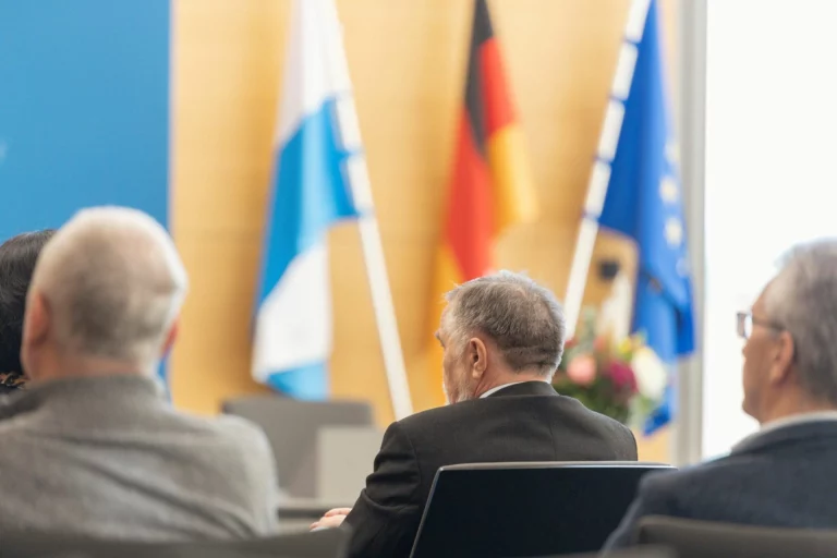 Mehrere Personen sitzen vor Bayern-, Deutschland- und Europaflagge bei einer Feierstunde