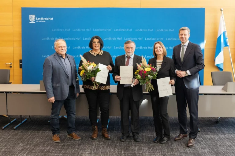 Fünf Personen stehen vor Landkreis-Hof-Wand, drei halten Urkunden und Blumensträuße