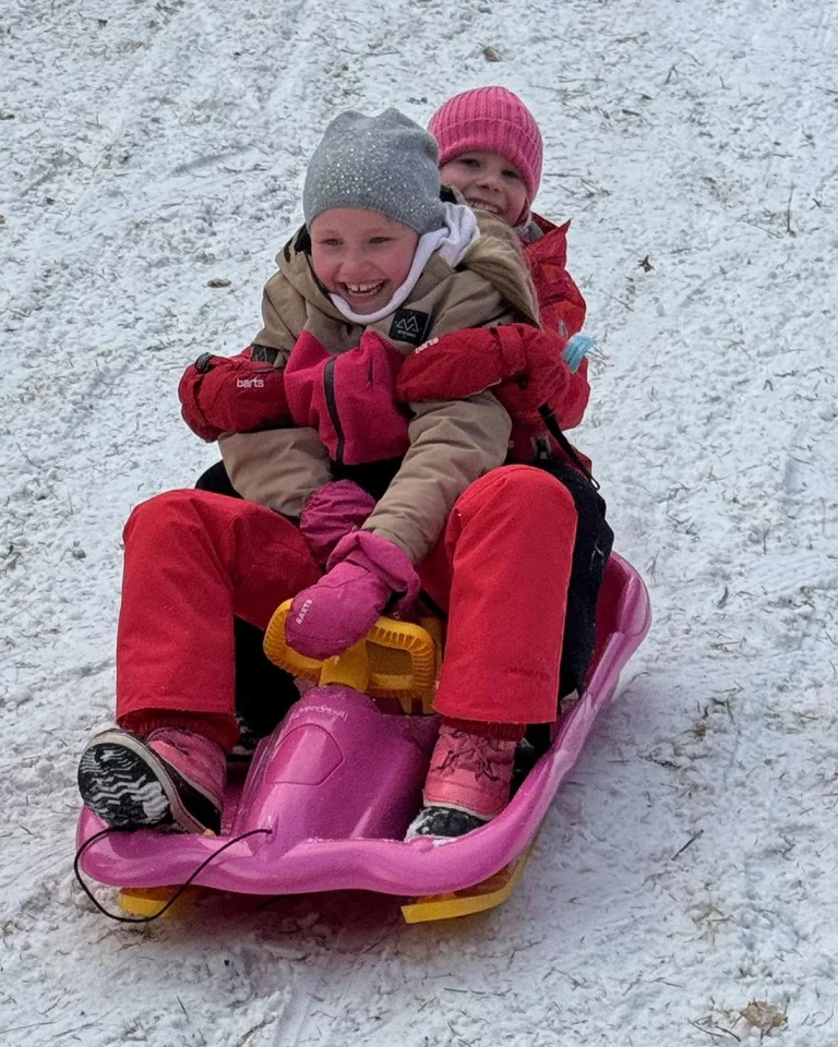 Zwei Kinder in Winterkleidung rodeln gemeinsam auf einem pinkfarbenen Schlitten den Hang hinunter