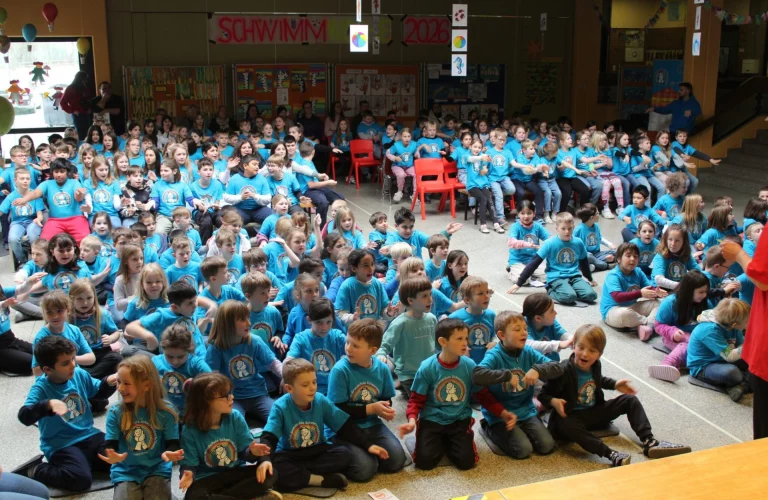 Große Gruppe von Grundschulkindern in türkisfarbenen Shirts sitzt und kniet vor einer Schwimmwoche-Dekoration