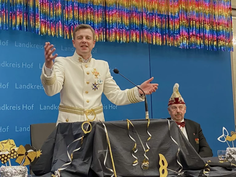 Mann in weißer Uniform mit Gardeorden am Rednerpult, daneben Person mit Narrenkappe