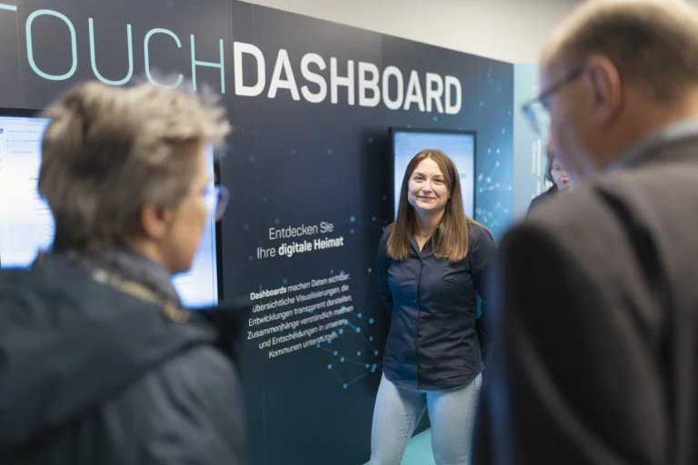Frau steht vor Touch-Dashboard-Stele, umringt von Besuchern im InnovationsRAUM