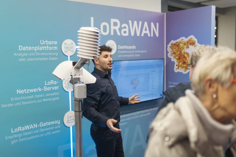 Mann erläutert LoRaWAN-Sensorik vor Ausstellungswand, im Vordergrund zwei Zuhörerinnen