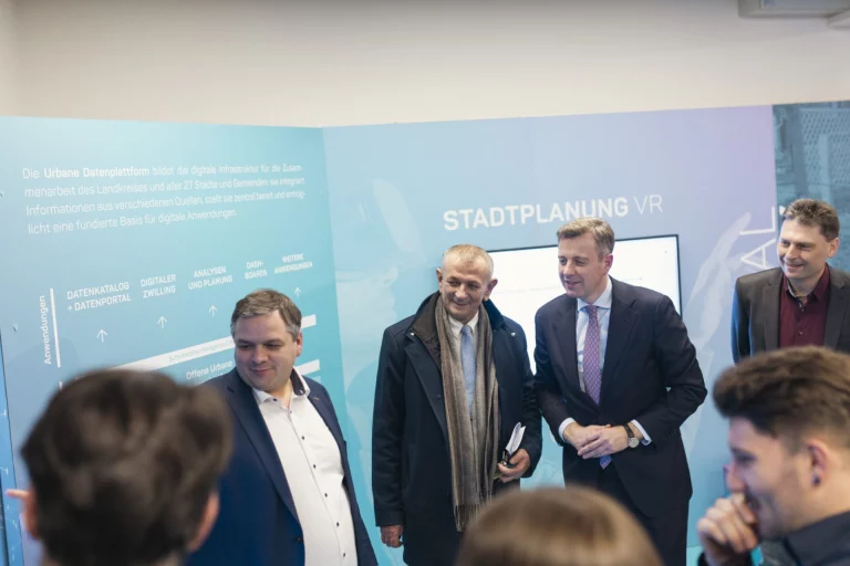 Männer in Anzügen vor Infotafel zur Urbanen Datenplattform mit Bildschirm „Stadtplanung VR“