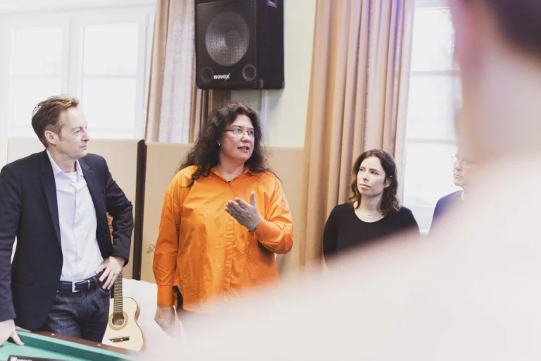 Mann, Frau mit erhobener Hand und weitere Personen im Gespräch neben Gitarren im Proberaum