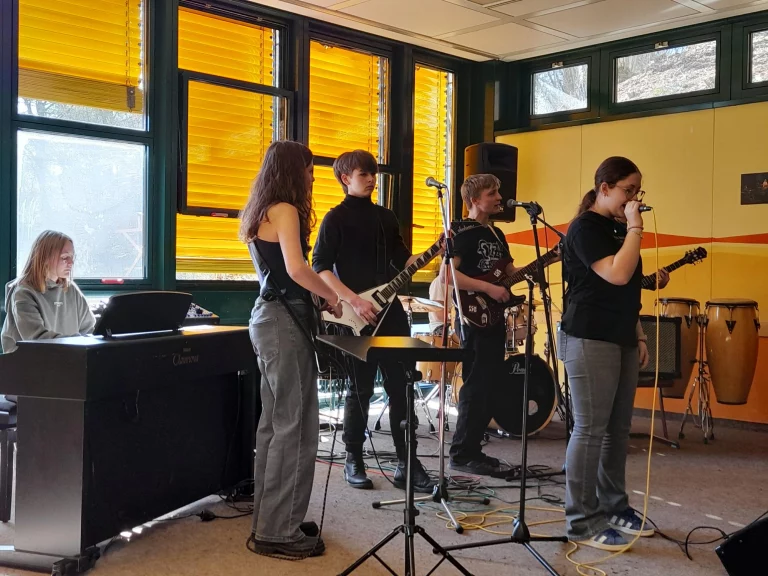 Fünf Jugendliche proben als Band mit E-Gitarren, Keyboard, Schlagzeug und Mikrofonen im Musikraum