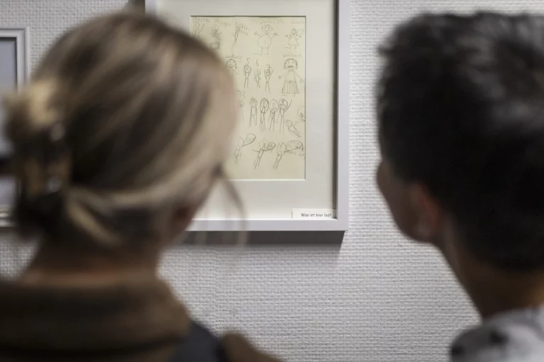 Zwei Personen betrachten gerahmte Kinderzeichnung mit Fragezeichen-Beschriftung in Ausstellung