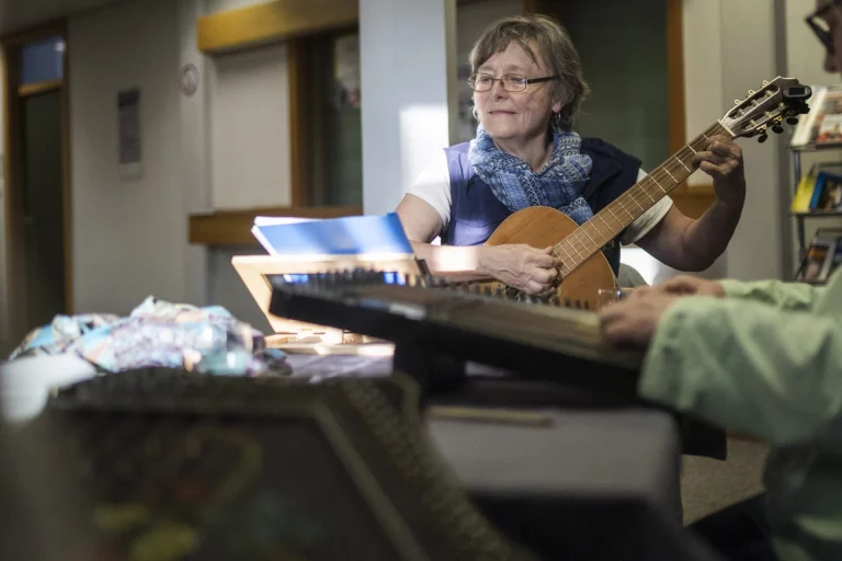 Frau spielt Gitarre neben Person mit Hackbrett, im Hintergrund Notenständer und Bücherregal