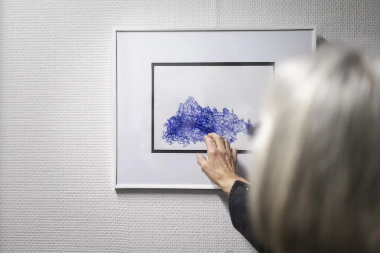 Hand an gerahmter Zeichnung mit dichtem blauen Linien- und Kritzelmuster an einer Ausstellungswand