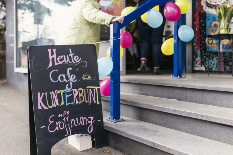 Tafel mit Aufschrift „Heute Café Kunterbunt – Eröffnung“ vor Treppe mit Luftballons