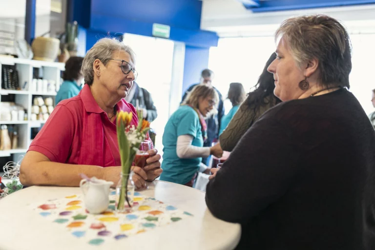 Zwei Frauen im Gespräch an einem Stehtisch mit Blumenvase, im Hintergrund weitere Gäste