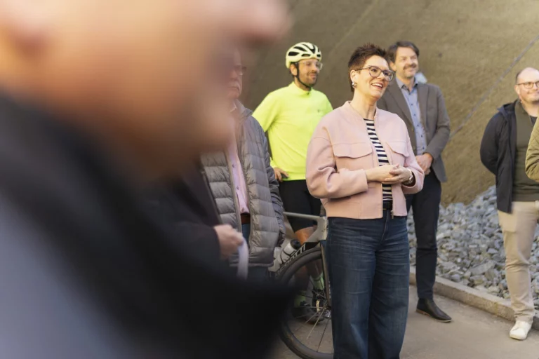 Lächelnde Frau in rosa Jacke bei der Einweihung im Kreis von Umstehenden, im Hintergrund ein Radfahrer