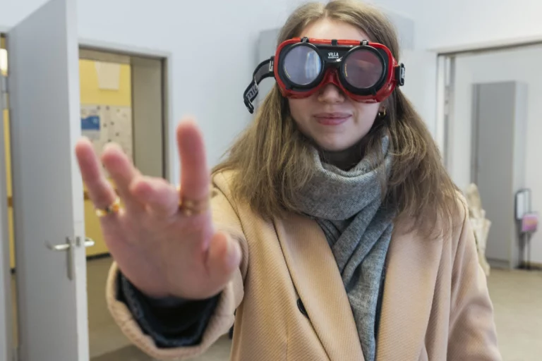 Frau mit roter Schutzbrille für Sehbeeinträchtigung macht Handzeichen in Fluren des Gesundheitsamts