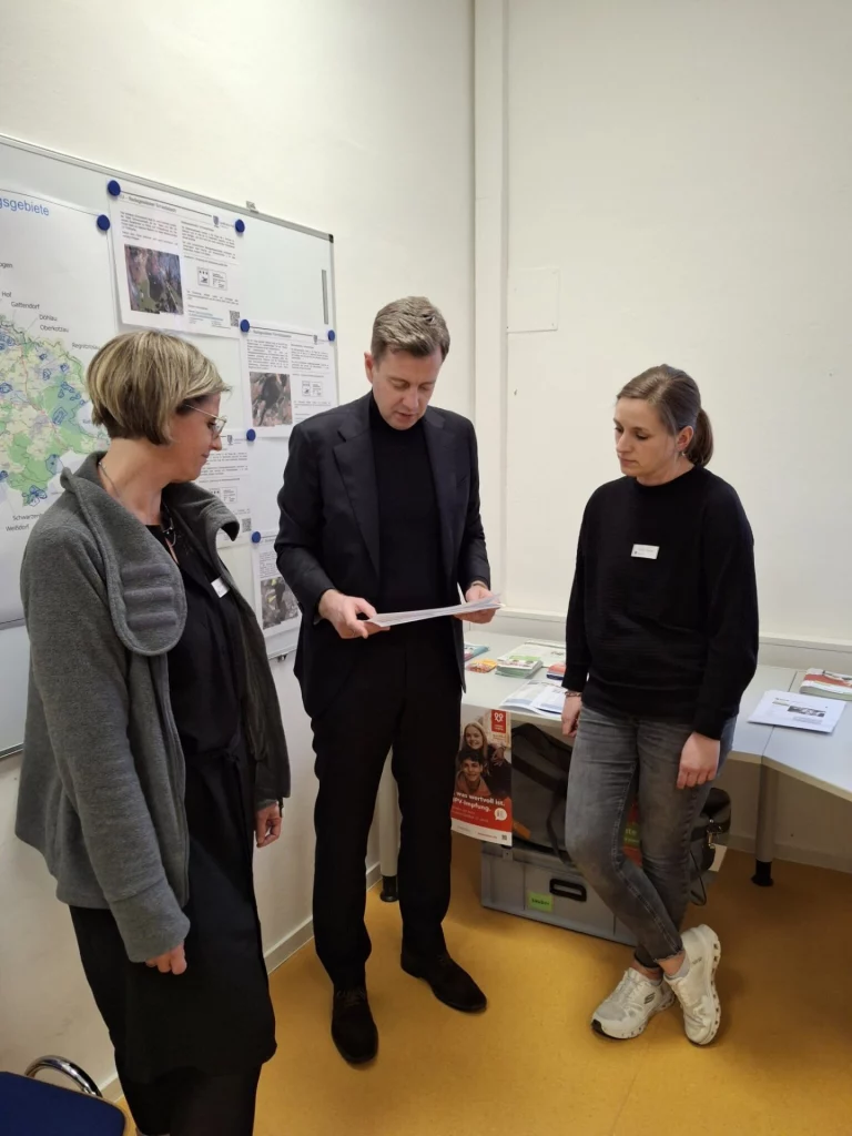 Mann im Anzug liest ein Dokument, zwei Frauen stehen daneben vor einer Infowand im Gesundheitsamt