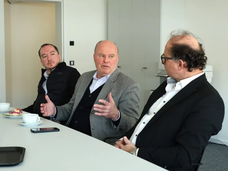Drei Männer im Gespräch am Konferenztisch, einer gestikuliert erklärend