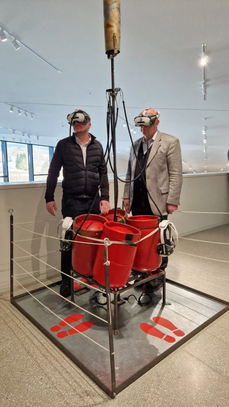 Zwei Männer mit VR-Headsets an einer Installation mit roten Behältern im Museum