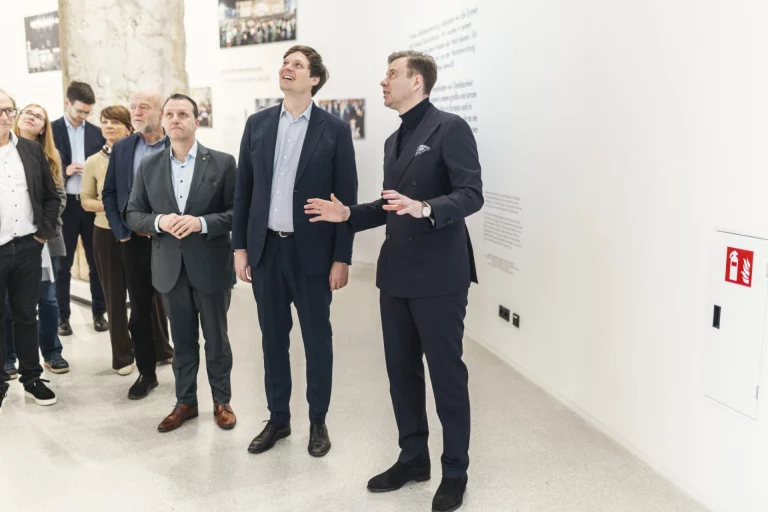 Gruppe von Personen in Ausstellungsraum, zwei Männer im Gespräch betrachten Exponate an der Wand