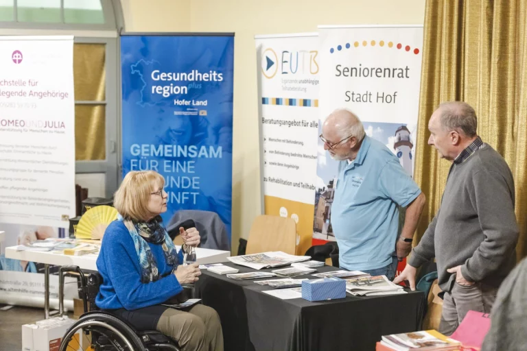 Informationsstand mit Rollups zu Gesundheitsregion und Seniorenrat, Frau im Rollstuhl im Gespräch mit zwei Männern