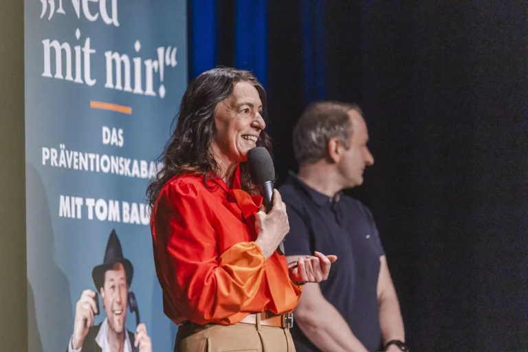 Frau mit Mikrofon spricht auf der Bühne vor Plakat „Ned mit mir!“, daneben ein Mann