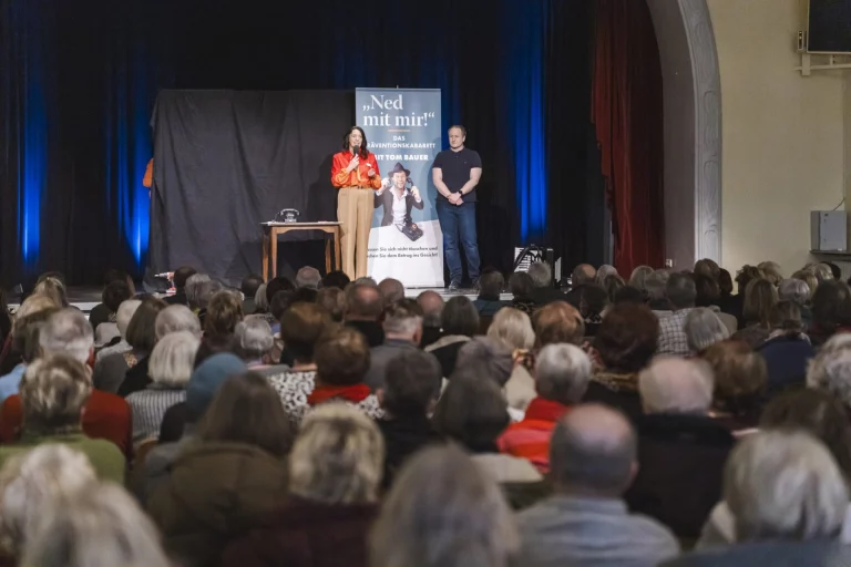 Zwei Personen auf der Bühne vor Plakat „Ned mit mir!“ und Publikum im vollen Saal