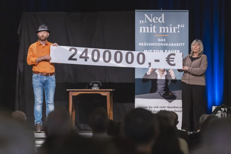 Mann und Frau halten auf der Bühne einen Scheck über 2.400.000 Euro vor Publikum
