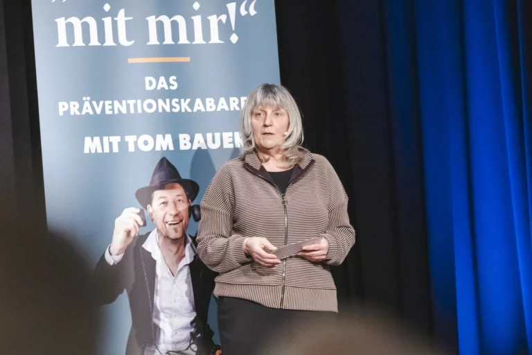 Frau mit Mikrofon auf Bühne neben Plakat des Präventionskabaretts „Ned mit mir!“ mit Tom Bauer