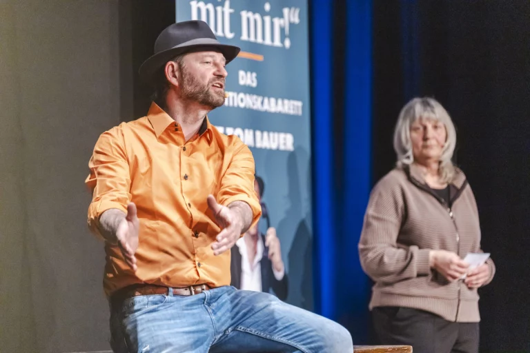 Mann in orangem Hemd gestikuliert auf der Bühne vor Plakat „…mit mir!“, Frau mit Zettel daneben