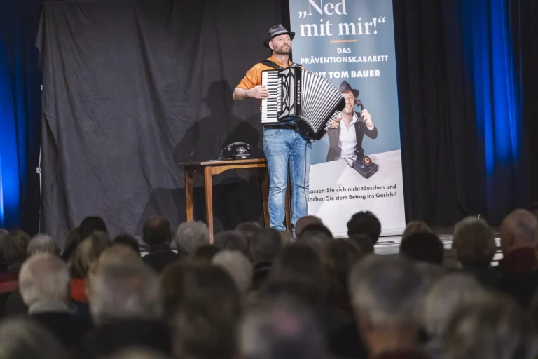 Mann mit Akkordeon auf der Bühne vor Publikum, daneben Plakat „Ned mit mir!“ zum Präventionskabarett