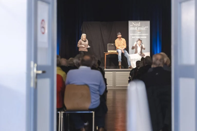 Zwei Personen auf einer Bühne vor Plakat „Ned mit mir!“, davor Publikum im Saal