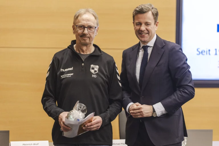 Mann mit Vereinstrikot und Pokal nimmt Auszeichnung von einem Mann im Anzug entgegen
