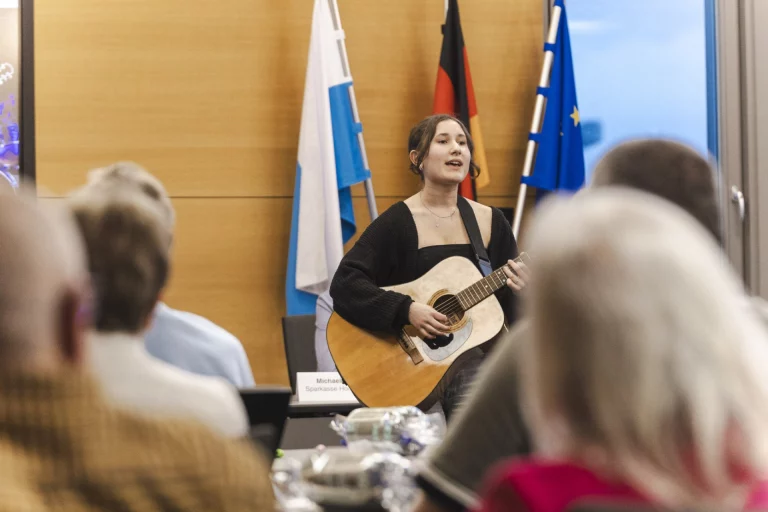 Junge Frau mit Gitarre singt vor Flaggen, Publikum im Vordergrund sitzend