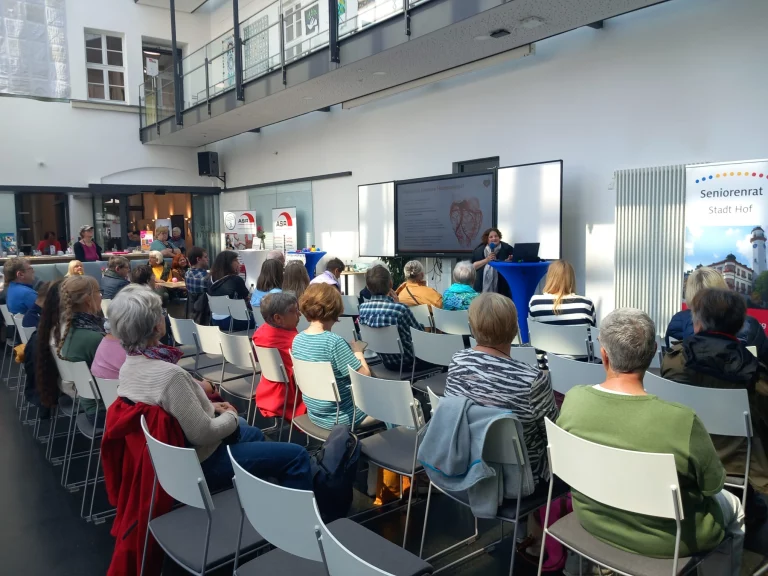 Zuhörerinnen und Zuhörer auf Stuhlreihen bei einem Vortrag, im Hintergrund Bildschirm mit Präsentation
