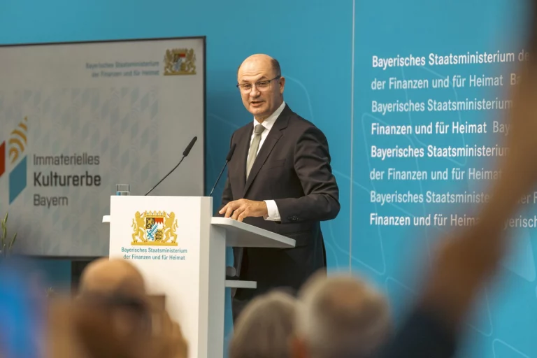 Mann im Anzug spricht am Rednerpult vor Publikum, Bühnenwand mit Schriftzug „Immaterielles Kulturerbe Bayern“