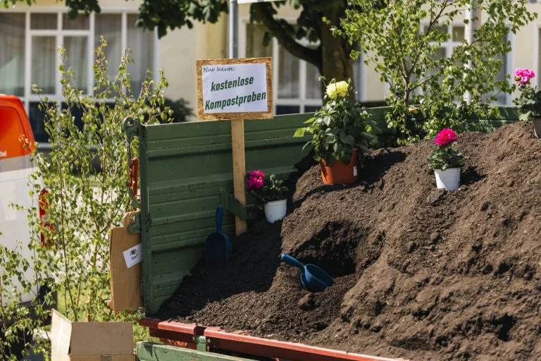 Schild „Nur heute: kostenlose Kompostproben“ an Container mit Komposthaufen und Blumentöpfen