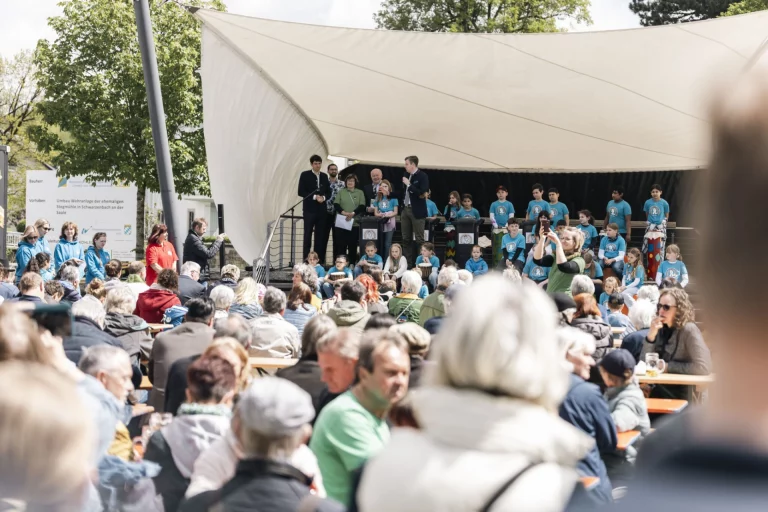 Menschenmenge bei Freiluftveranstaltung mit Bühne, Rednern und Kindergruppe in blauen T-Shirts