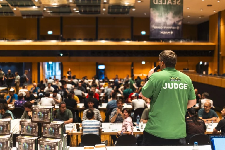 Judge spricht mit Mikrofon vor Teilnehmenden an Turniertischen in gefülltem Saal