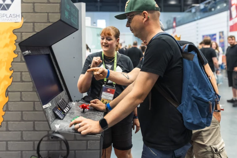 Besucher testet Arcade-Automat am Messestand, Standbetreuerin erklärt die Steuerung