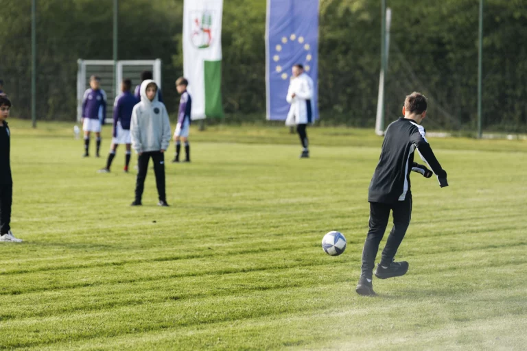 Jugendlicher schießt beim Training auf dem Rasen einen Fußball, im Hintergrund Flaggen und weitere Spieler