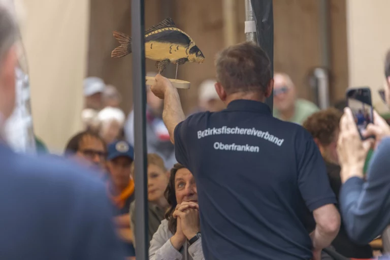Mann im Trikot des Bezirksfischereiverbandes Oberfranken hält Fischmodell hoch vor Publikum im Festzelt