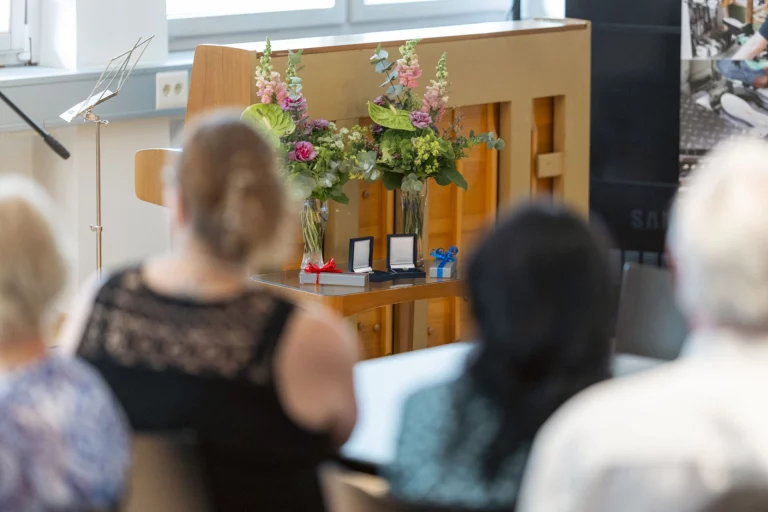 Blumensträuße und zwei Ehrennadeln auf einem Rednerpult, davor sitzendes Publikum