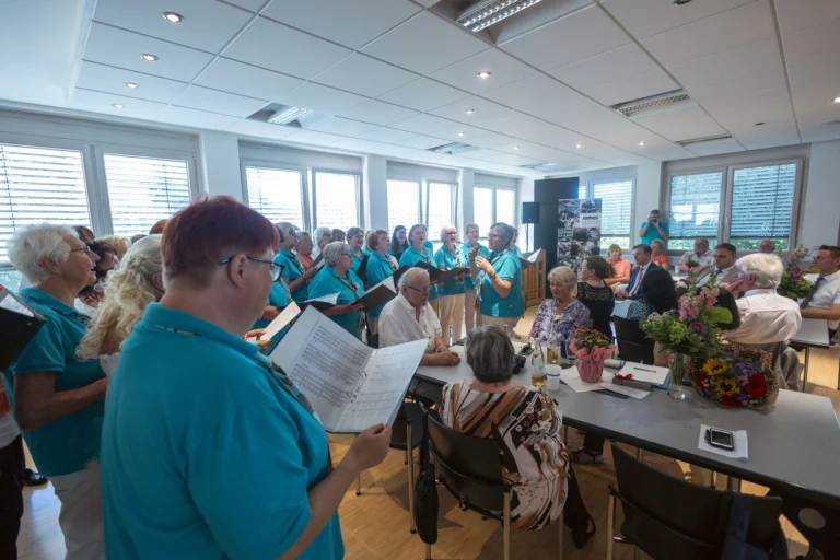 Chor in türkisfarbenen Shirts singt bei der Ehrung im Sitzungssaal, davor Tisch mit Blumensträußen