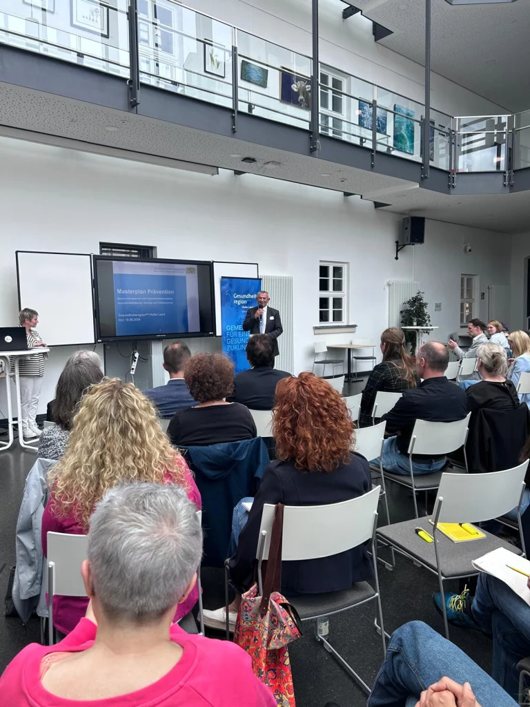 Mann am Stehpult spricht vor Sitzreihen, im Hintergrund Präsentation zum Masterplan Prävention