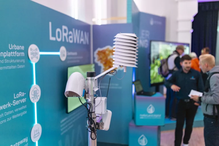 Messstation mit Wetter- und LoRaWAN-Sensoren vor Ausstellungswänden zur Datenplattform, im Hintergrund Besucher im Gespräch