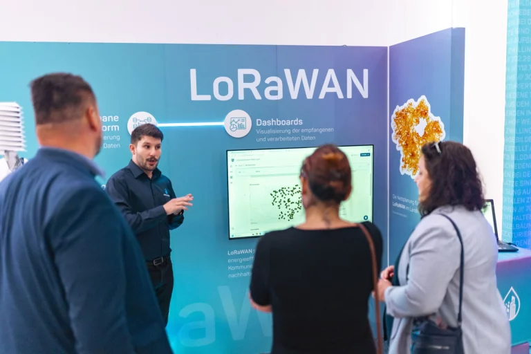 Mann erklärt Besuchergruppe eine LoRaWAN-Dashboard-Präsentation am Messestand